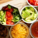Ingredients for a taco or taco salad arranged in small bowls on a wooden surface, including corn chips, chopped tomatoes, diced avocado, shredded lettuce, seasoned ground beef, shredded cheese, and salsa.