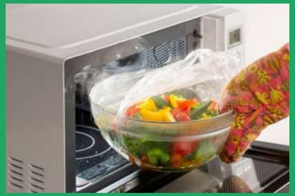 A microwave with fresh vegetables being put in to cook.