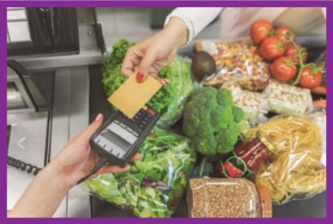 Person paying with a card at a grocery checkout while fresh produce and packaged foods sit on the counter.