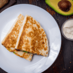 Two folded quesadillas on a white plate with an avocado half, sour cream, and seasoning on a dark wooden surface.