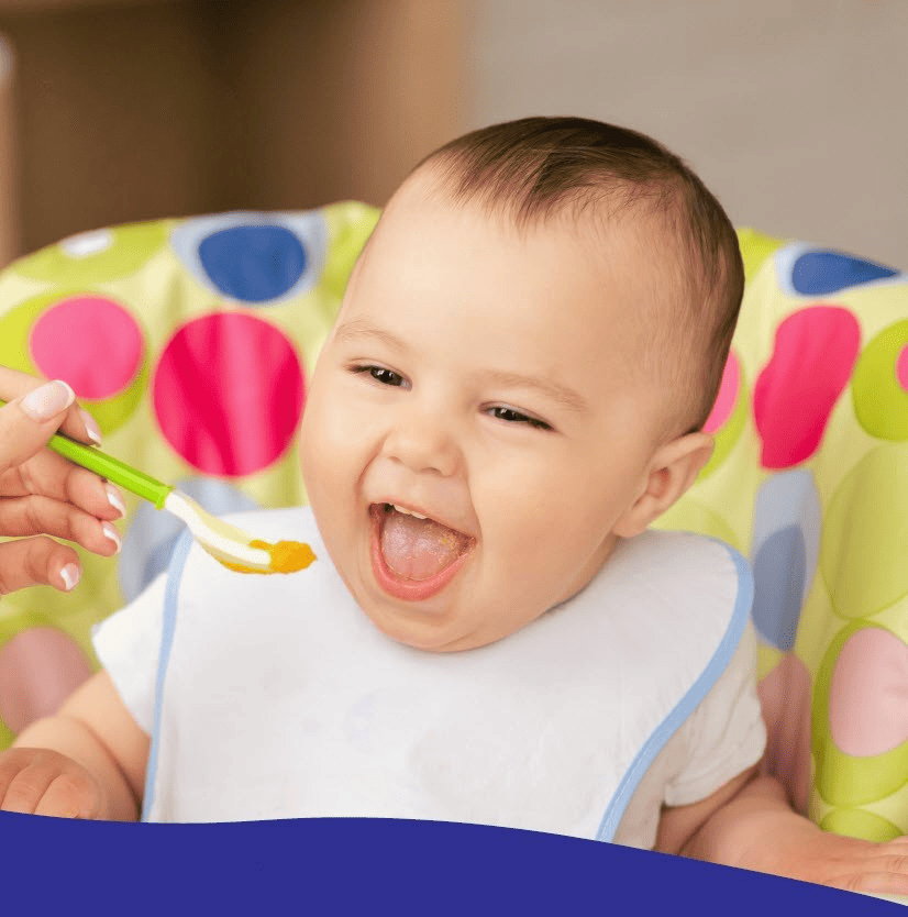 Baby sitting in a high chair with a bib, opening their mouth as an adult holds a spoonful of orange baby food toward them.
